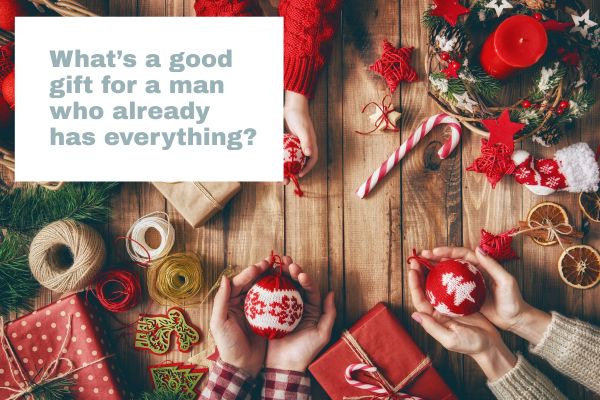 Festive flat lay with Christmas decorations, gift boxes, and hands holding ornaments beside a text box asking: “What’s a good gift for a man who already has everything?”